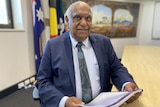 A senior Aboriginal man wearing a suit and tie sits at a table holding a document while smiling at the camera