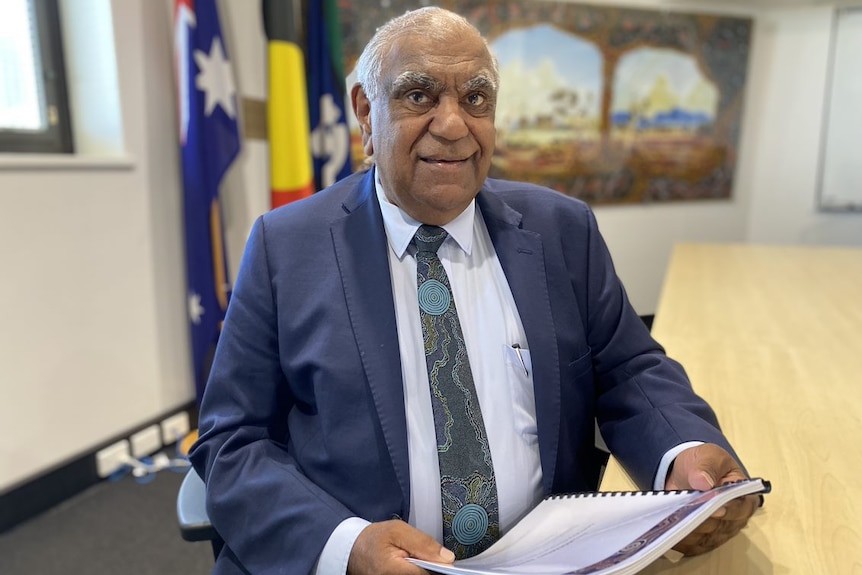 A senior Aboriginal man wearing a suit and tie sits at a table holding a document while smiling at the camera