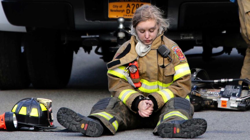 Female firefighter with black on her face sits on the ground behind a truck while two male firefighters lean against it.