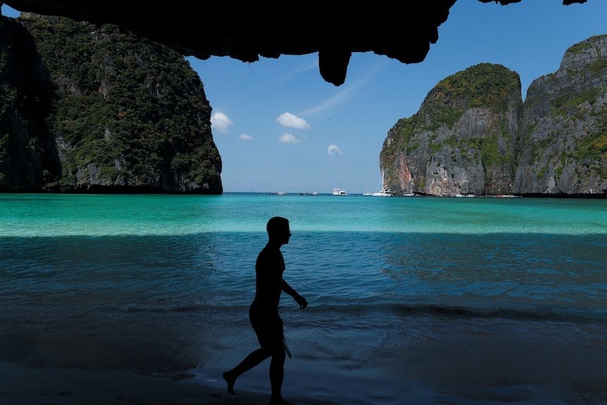 A silhouette walks along a beach in front of a bright blue sea, surrounded by cliffs. 