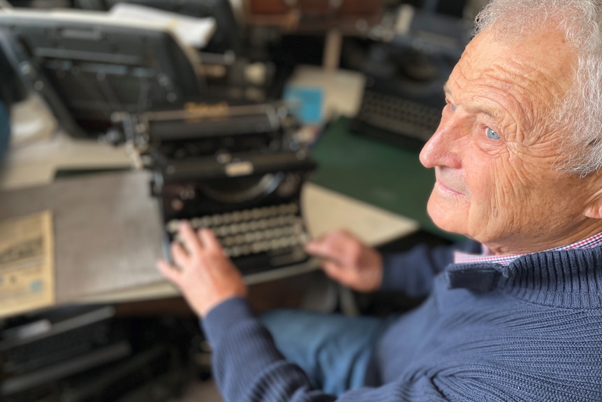 An elderly man sits at a desk, his hands resting on the keys of a typewriter.