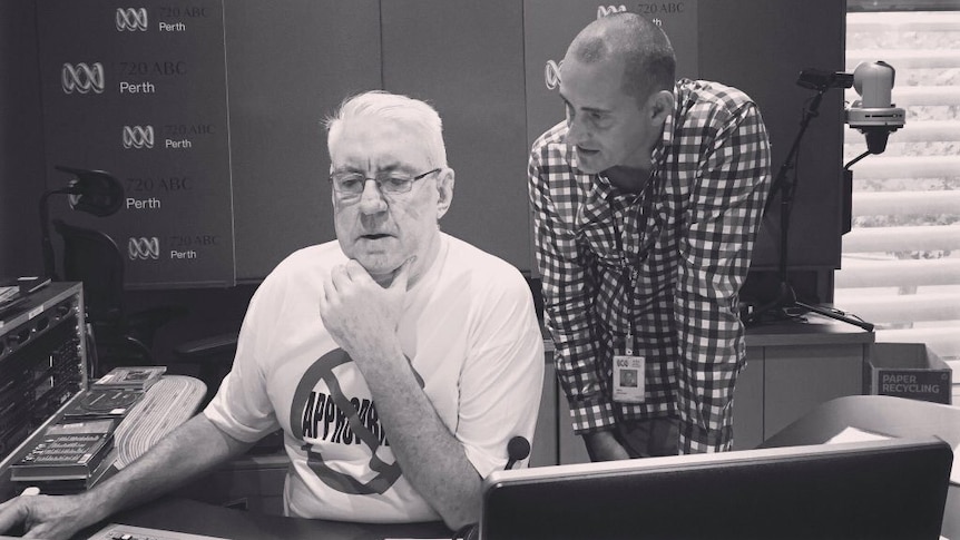 A black and white shot of Eoin Cameron sitting in a radio studio behind a desk looking at a computer with producer Brad McCahon