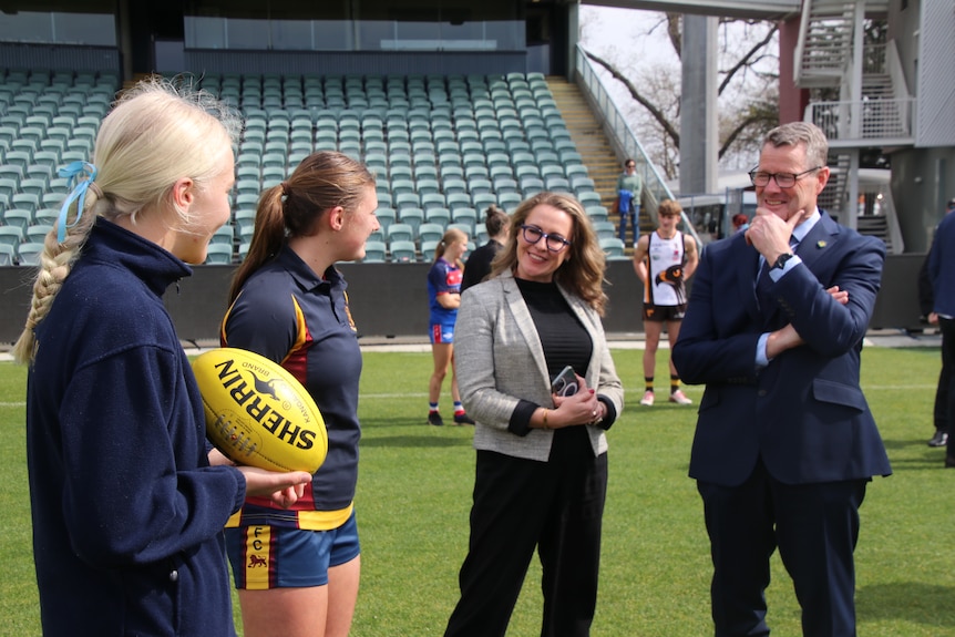 Two young female football players speak to a businesswoman and a man in a suit on a football oval