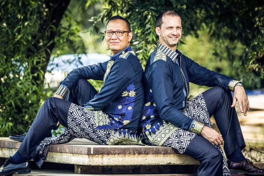 Two men dressed in matching dark blue pants and top sit back to back on a ledge.