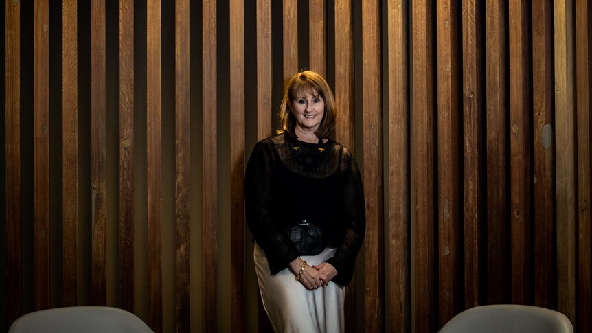 Kirsten O'Doherty stands against a wood panel background with a light overhead. She is smiling and wears a black shirt.