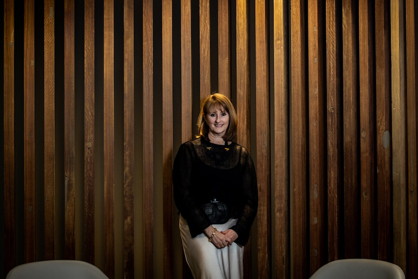 Kirsten O'Doherty stands against a wood panel background with a light overhead. She is smiling and wears a black shirt.