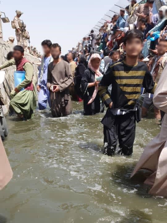 A group of Afghan people stand in flood waters at Kabul airport, their faces blurred for privacy. 