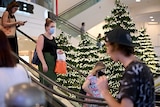 A mask-wearing woman holds a shopping bag as she descends an escalator at Myer's Sydney store, surrounded by Christmas trees.