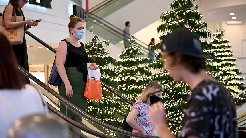 A mask-wearing woman holds a shopping bag as she descends an escalator at Myer's Sydney store, surrounded by Christmas trees.