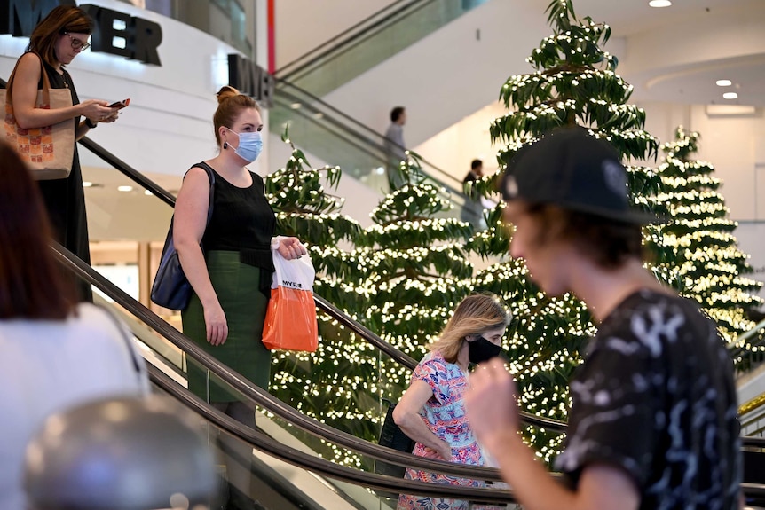 A mask-wearing woman holds a shopping bad as she descends an escalator at Myer's Sydney store, surrounded by Christmas trees.