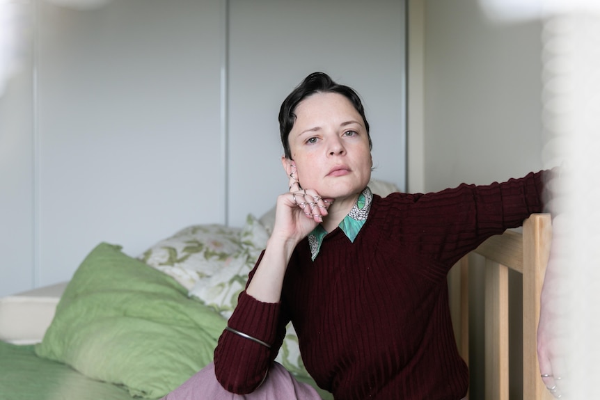 A white woman in her mid-30s with short brunette hair sits on a bed with green bedding