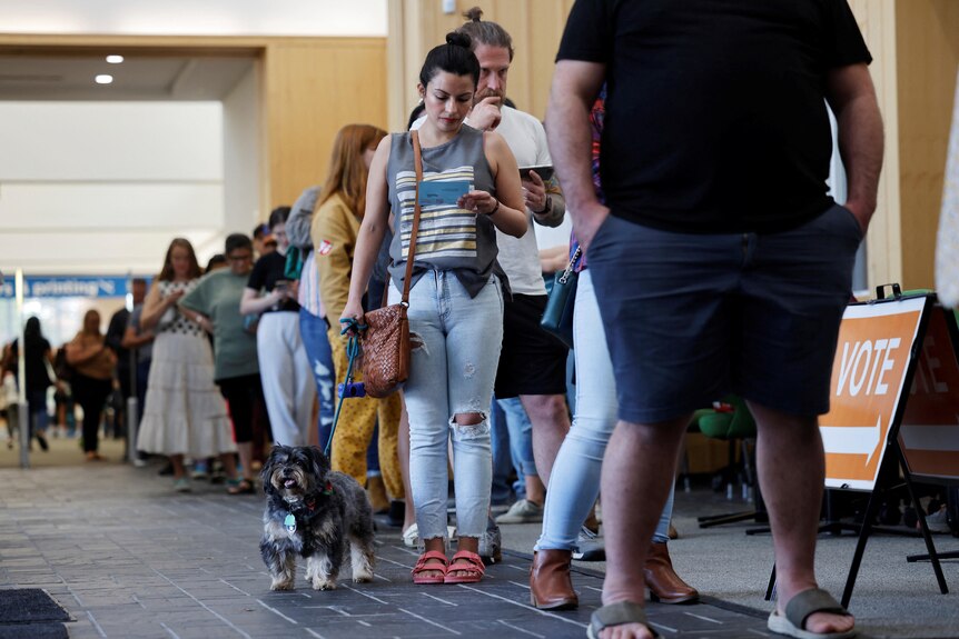A woman holding a little dog on a leash stands in a long line 