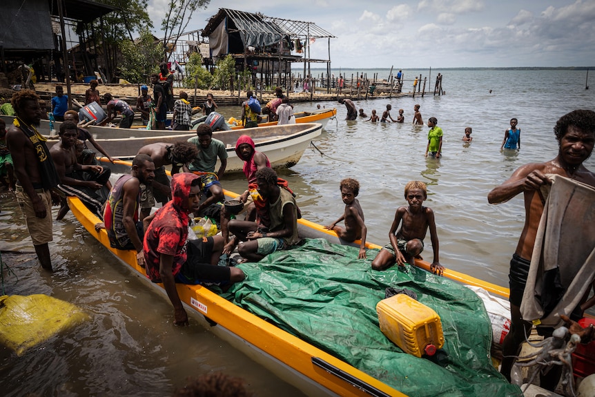 Children wade in the shallow water near two boats on the shore, filled with people
