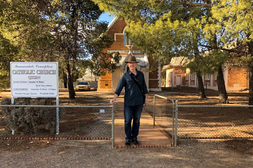 A tall man in jeans, a navy shirt and Akubra-style hat stands at the front gate outside a Catholic church in Quorn