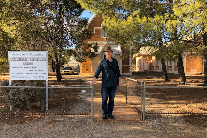 A tall man in jeans, a navy shirt and Akubra-style hat stands at the front gate outside a Catholic church in Quorn