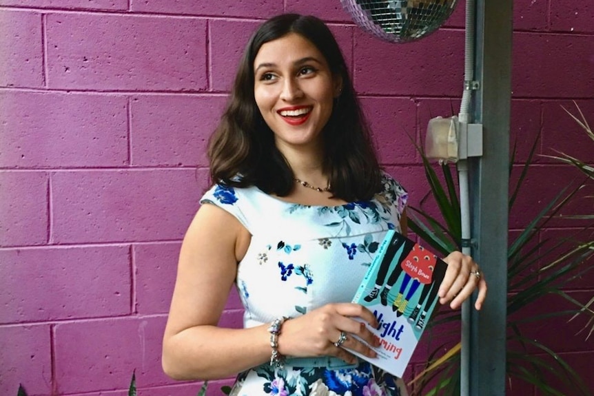 Steph Bowe pictured holding a book with a pink wall and disco ball in the background, pictured in obituary for Steph Bowe