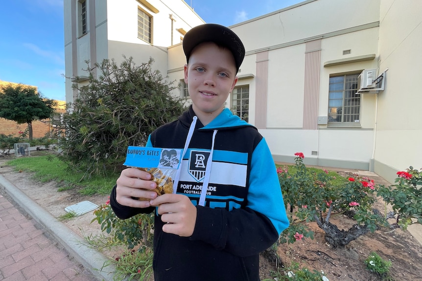 A boy in a port adelaide jumper holds a plastic bag which has Buddy's Bites written on it, the bag contains small biscuits