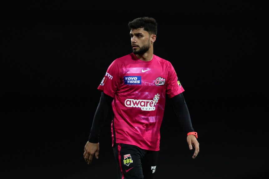 Sydney Sixers player Naveen-ul-Haq walks across the field with a dark background behind him.