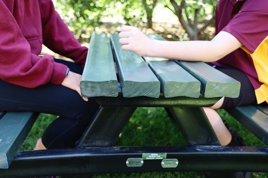 Kids in maroon and yellow uniforms sit at a green plastic picnic table