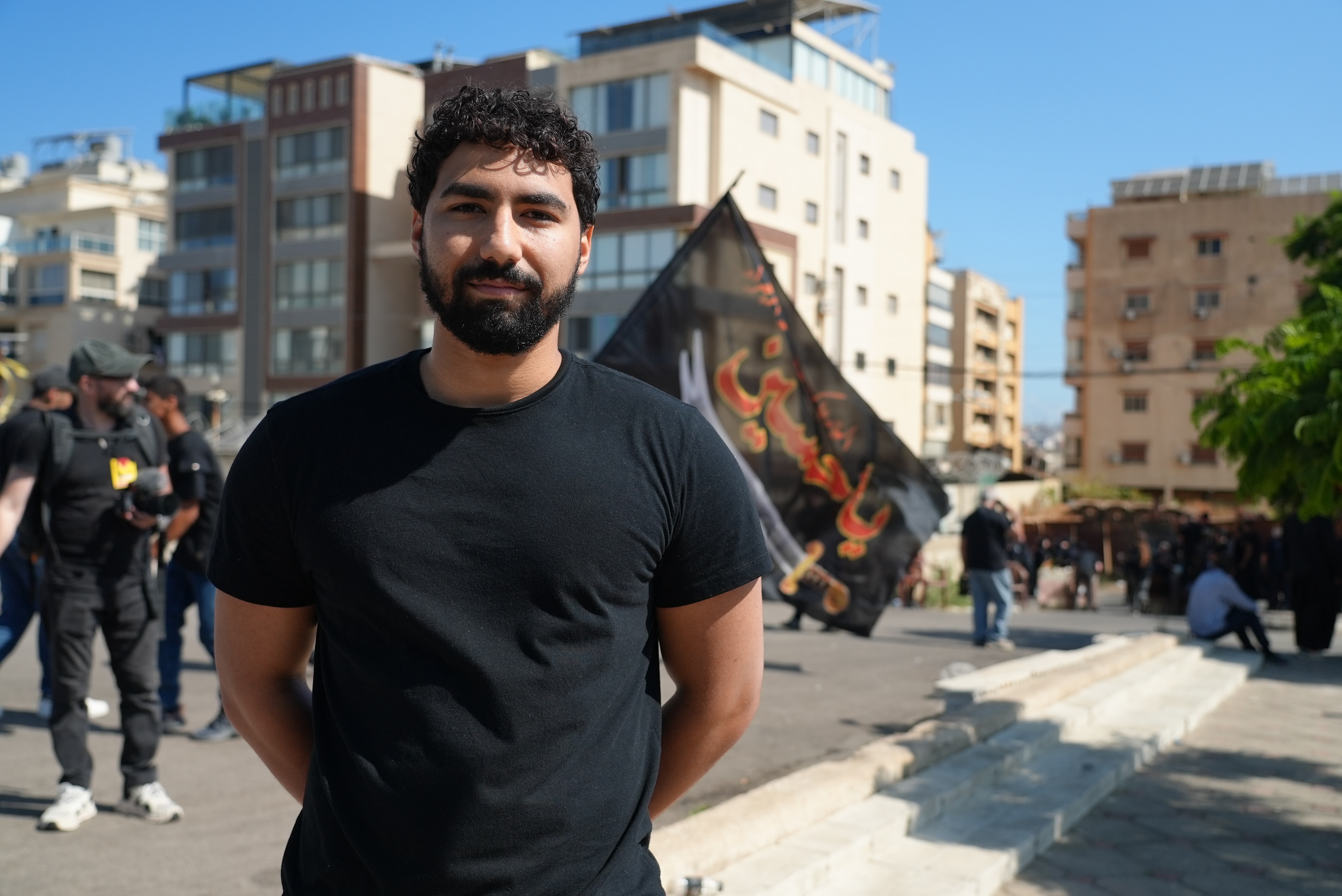 A man in his 20s, with short curly black hair, a black beard, wearing a black T-shirt.