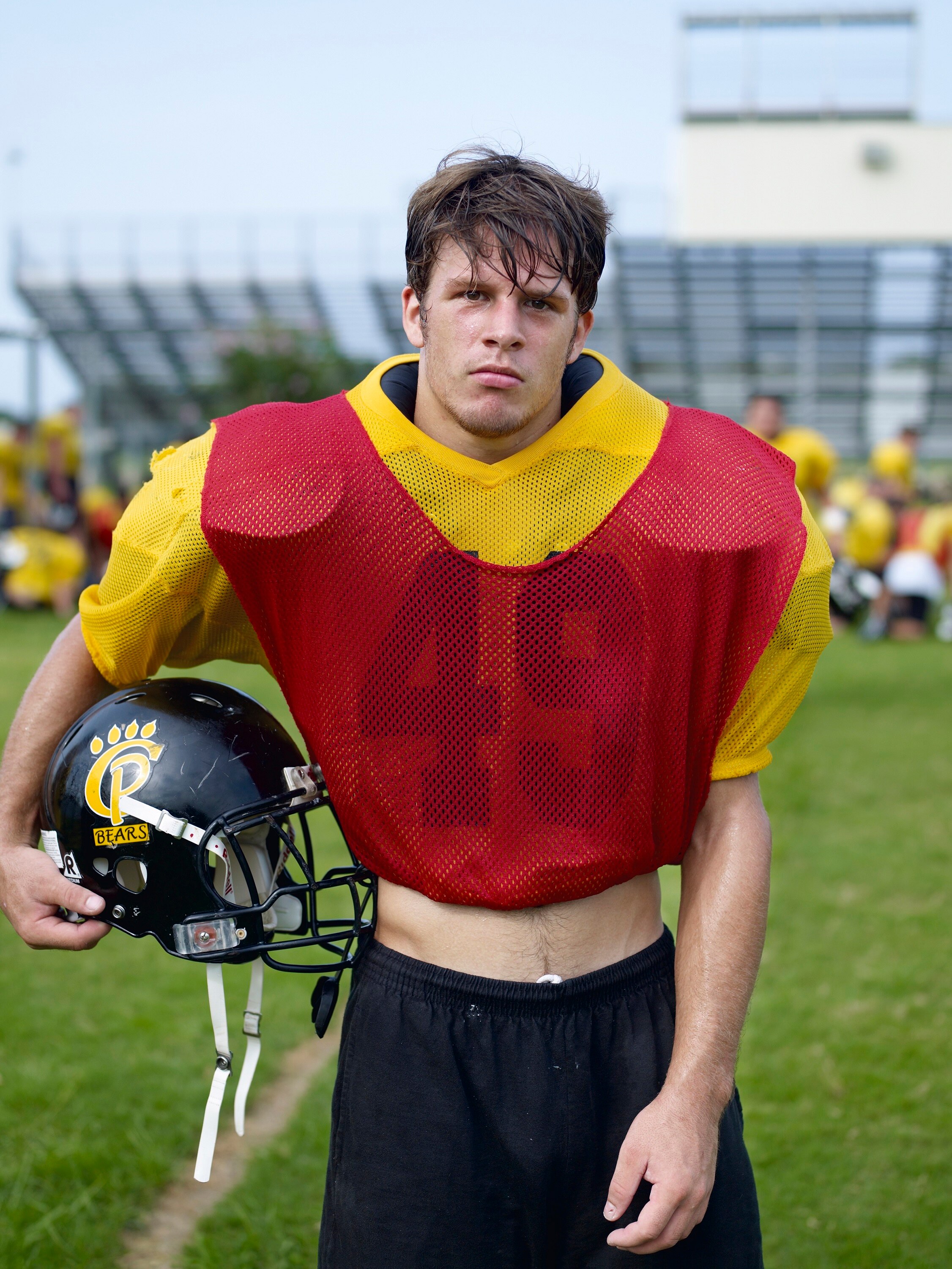 A young white man wears a red and yellow grid iron uniform, holding a black helmet at his side