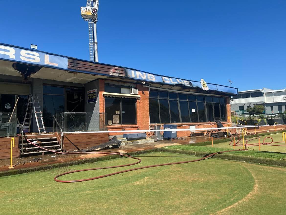Las mangueras contra incendios yacen sobre la hierba verde frente a un edificio de ladrillo con un letrero azul que dice RSL que fue quemado por el fuego.