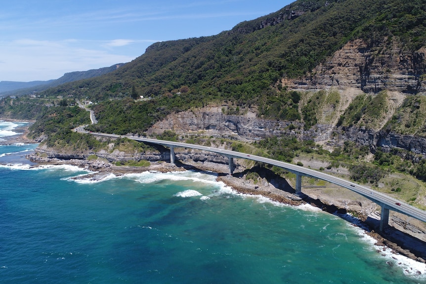 The Sea Cliff Bridge as seen from a drone, looking south towards Clifton with blues skies and blue water below.