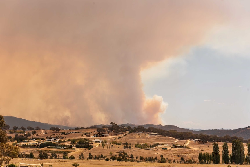 A plume of smoke rises up from behind a hill, houses dotted about nearby.