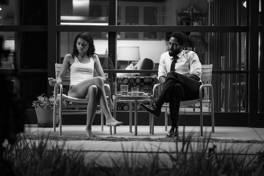 Black and white photo of young man and woman sitting on chairs on patio, him looking at her as she looks away.