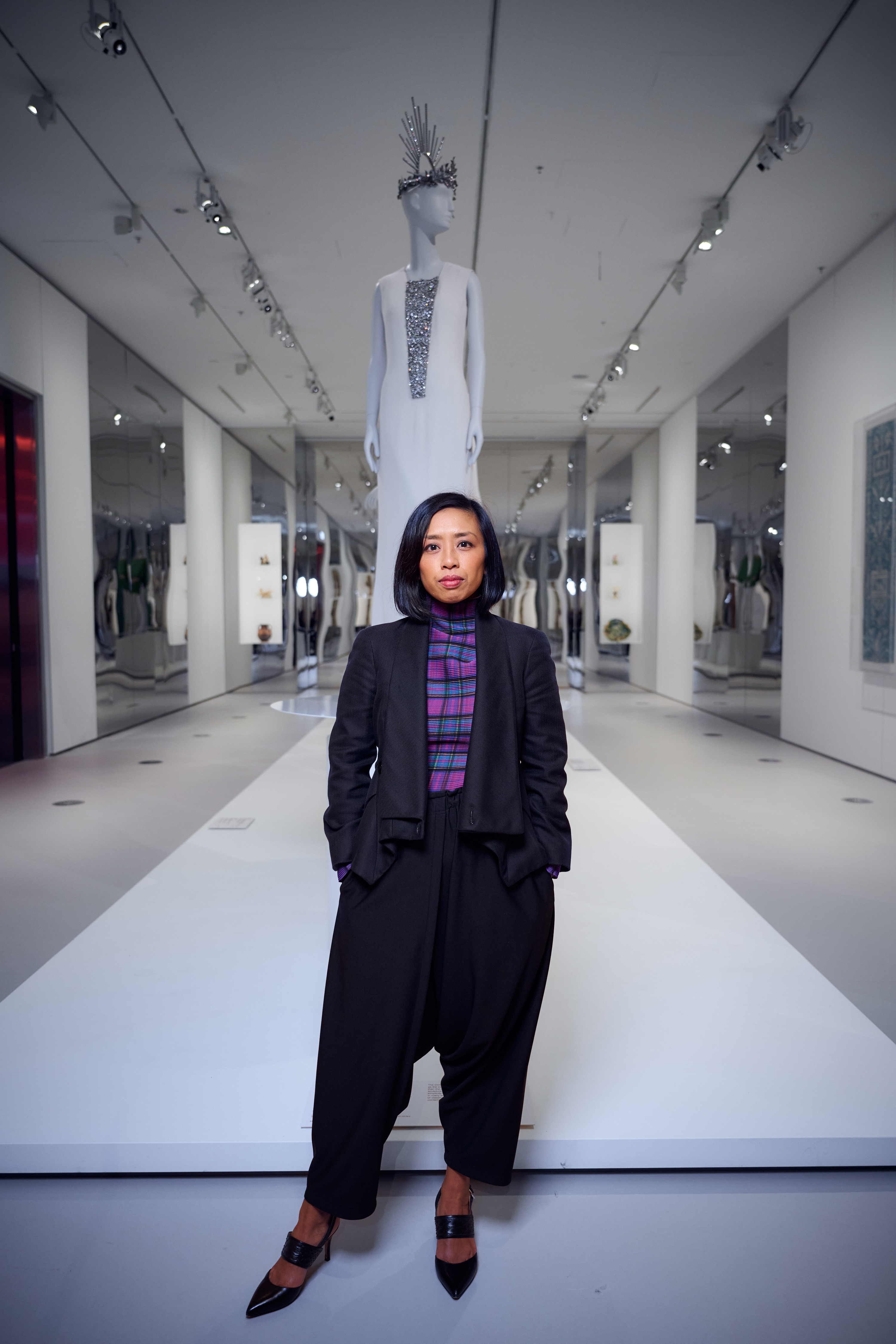 Asian woman with short dark hair wears a vibrant checkered purple shirt under a sleek black coat and pants in a gallery.