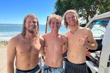 three men smiling shirtless in front of beach