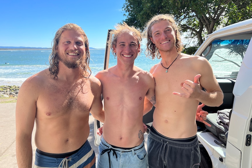 three men smiling shirtless in front of beach