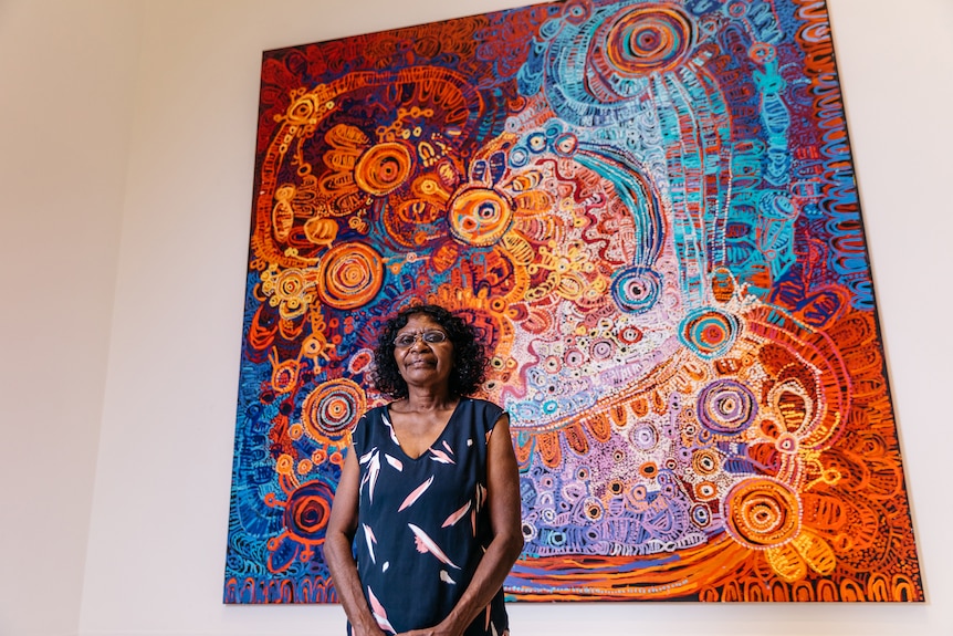 Pitjantjatjara woman with curly brown hair and glasses wears navy dress and stands in front of richly coloured painting.