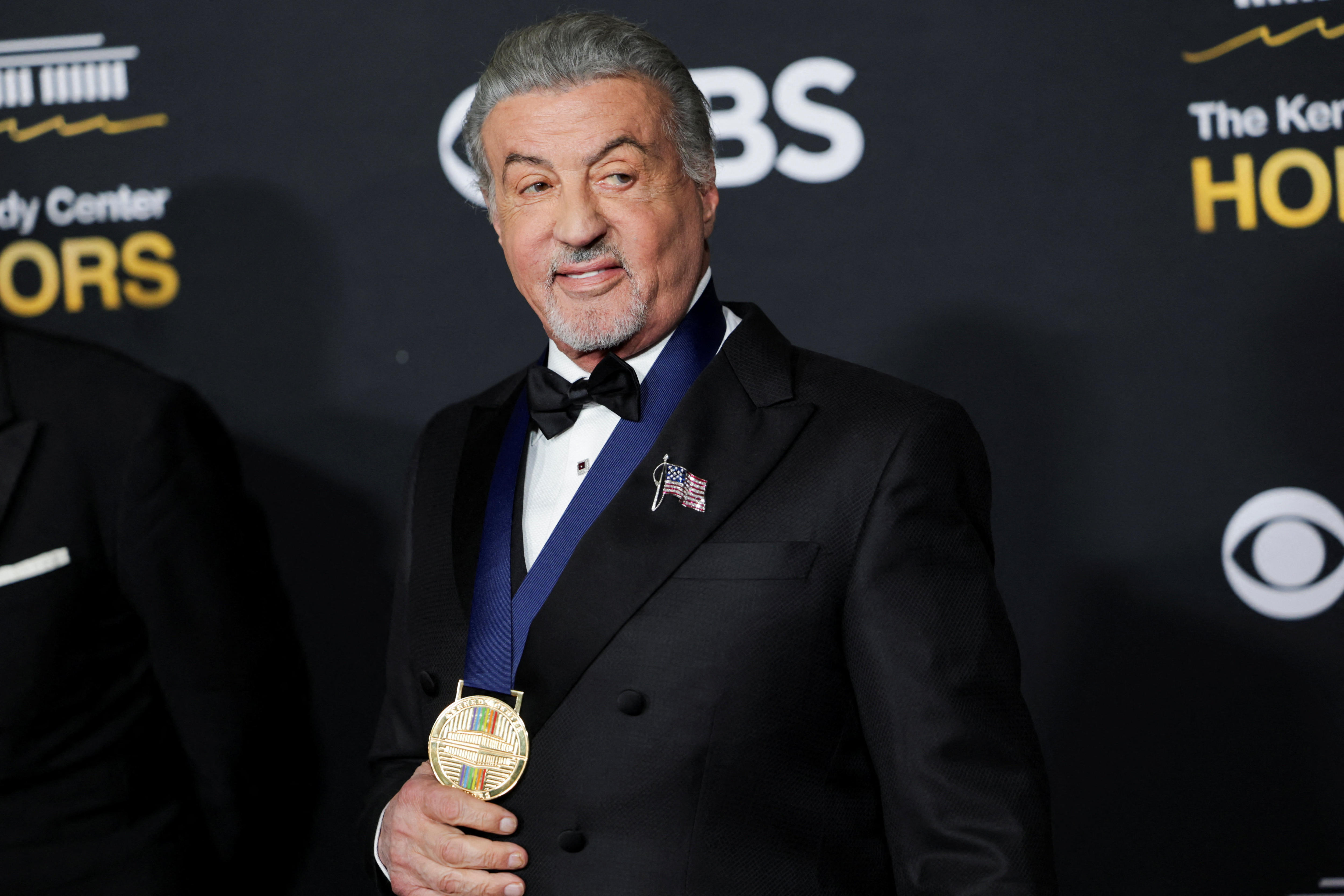 Sylvester Stallone viste un esmoquin negro sobre un fondo oscuro y lleva una medalla con una cinta azul alrededor del cuello.