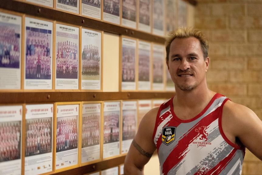 Man in red and white jersey smiles in front of a wall filled with sport team photos.