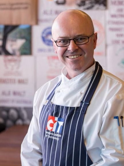 A man in an apron wearing glasses smiles, standing in a cafe