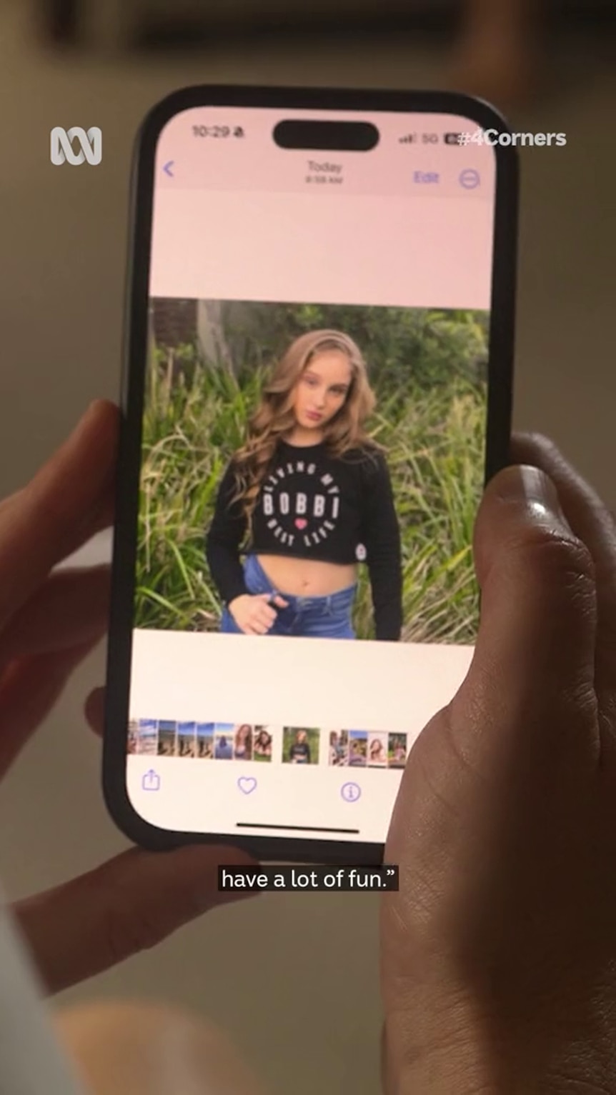 A white person's hand holds a smartphone showing a white girl posing in front of some greenery 