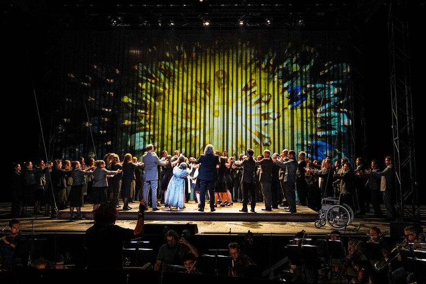 A large cast of opera singers are congregated on stage in a circle with their arms extended towards its centre.