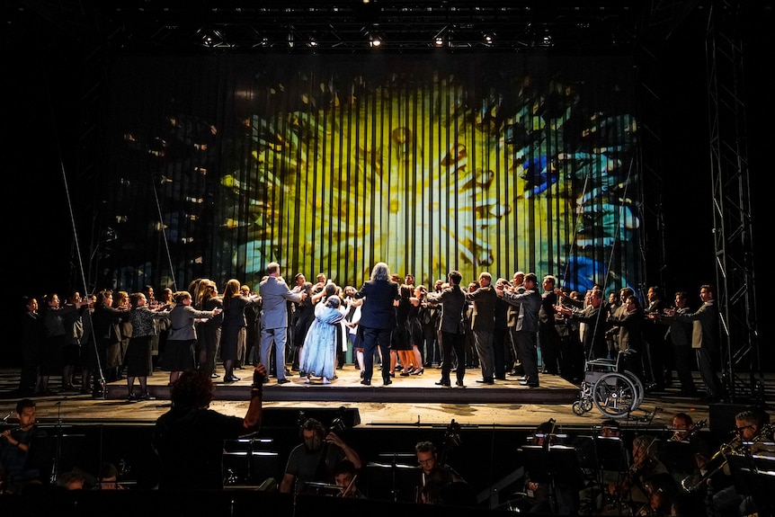 A large cast of opera singers are congregated on stage in a circle with their arms extended towards its centre.