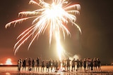 Two dozen people line a beach watching a firework explode overhead.