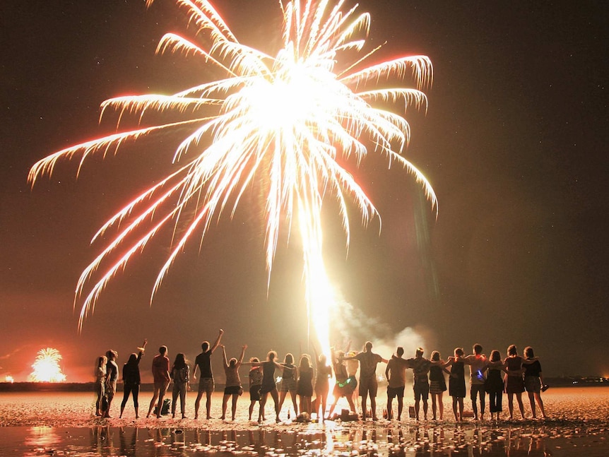Two dozen people line a beach watching a firework explode overhead.