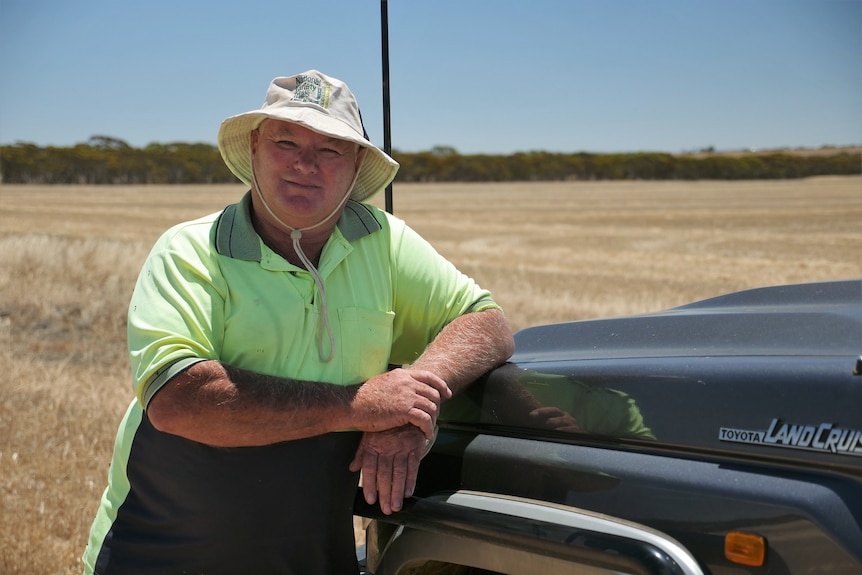 A man wearing a reflective T-shirt and a white hat leaning against car bonnet in barley field.