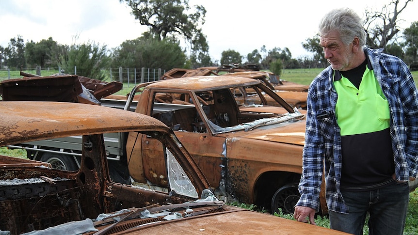 A man stands in front of seven burned-out, rusted car wrecks.