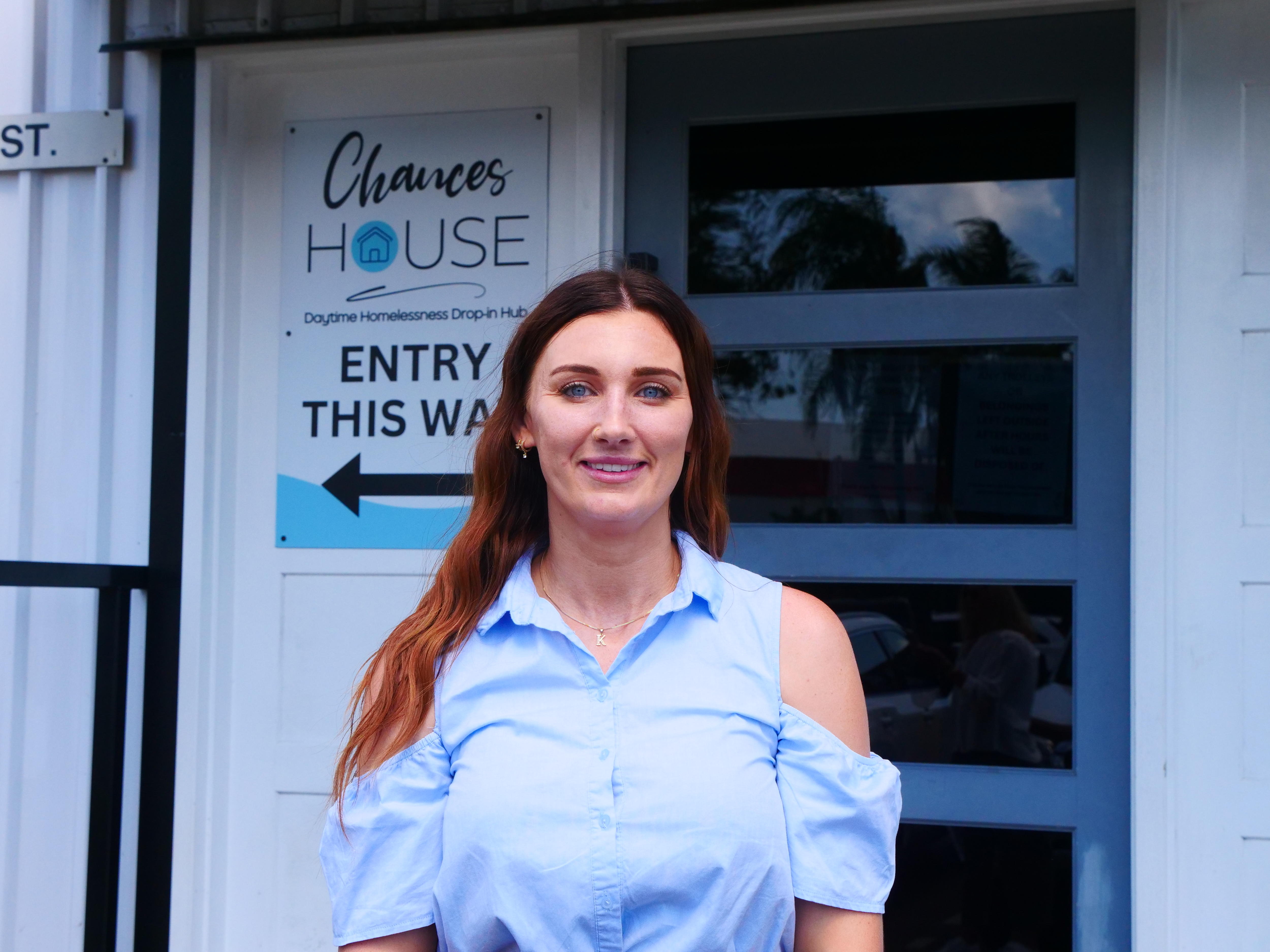 Uma mulher está do lado de fora de um abrigo para moradores de rua. Uma placa com uma seta diz "entrada aqui". 