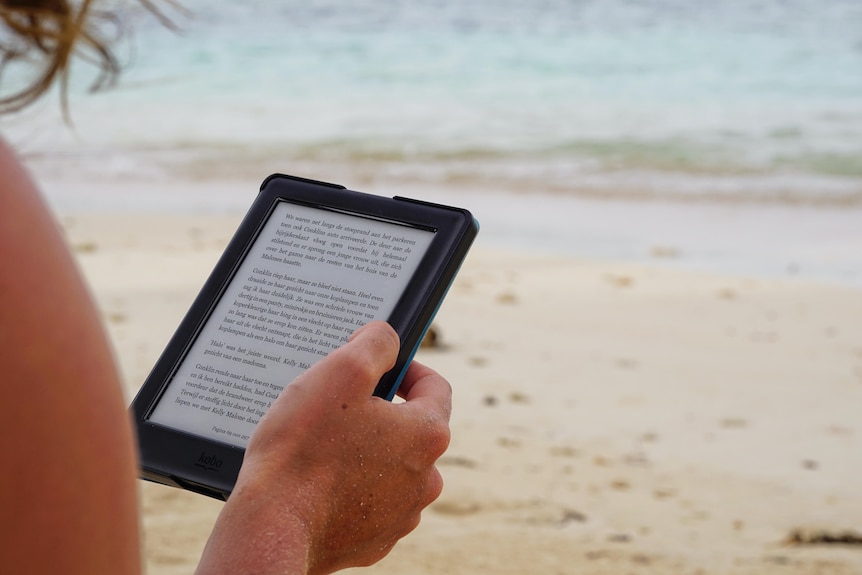 A hand holding an e-reader with sand and water in the background.