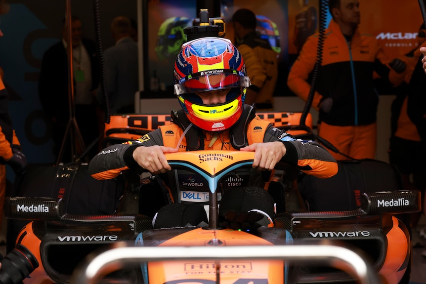 An F1 driver in orange lowers himself into his car, which is in the garage