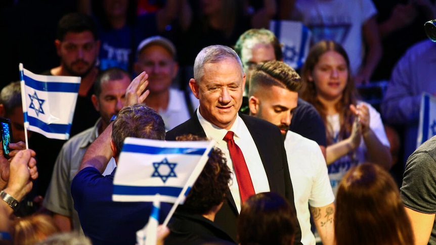 Benny Gantz walks on stage surrounded by a large crowd of people waving Israeli flags.