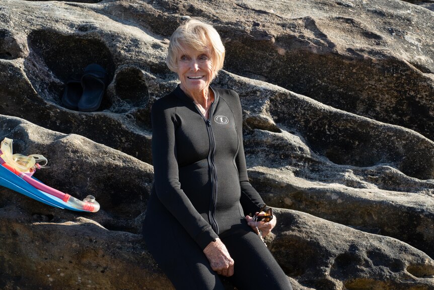 Elderly woman in black wetsuit smiles sitting on some rocks, with a snorkel set next to her