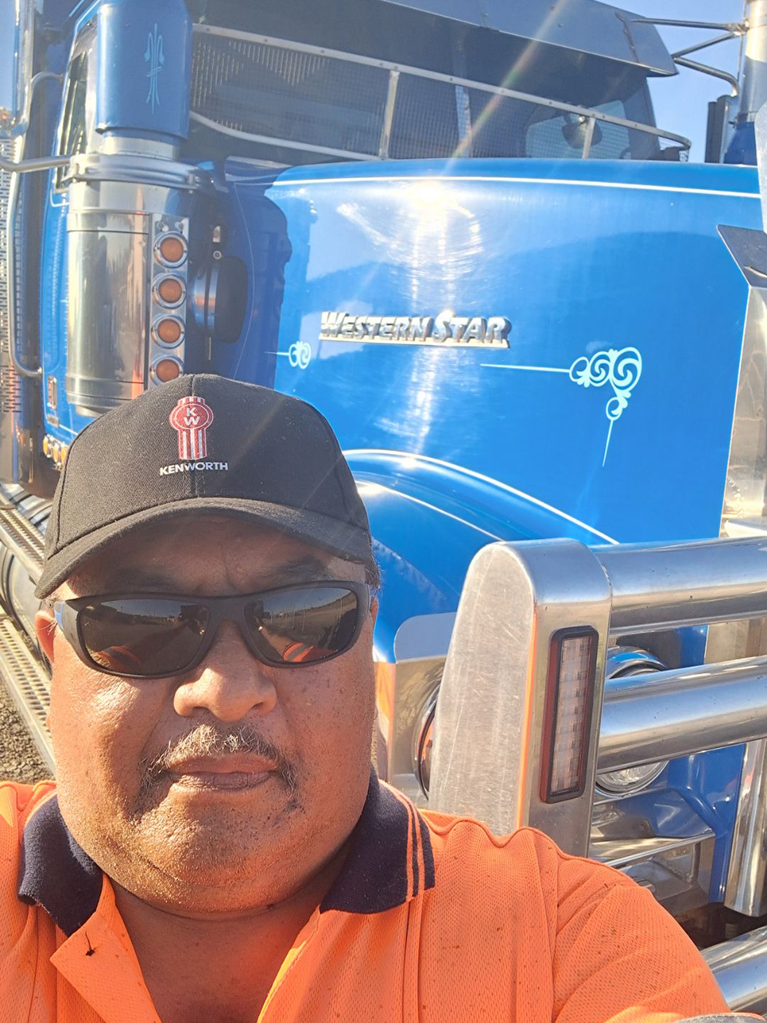 Una selfie de Johnnie Tumai con gorra y gafas de sol parado frente a una camioneta azul. 