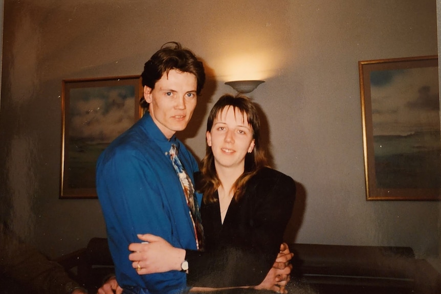 An old coloured photo of a man in a blue shirt, tie, holding a girl in black, paintings behind them.